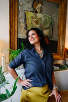 A woman posing in a navy blue button-down shirt and a mustard yellow skirt, standing in front of a large framed painting in an indoor setting.