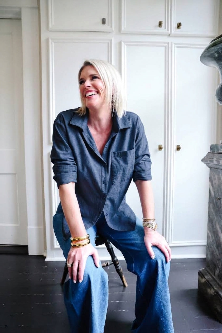 A smiling blonde woman sits on a stool in a room with white cabinets, wearing a dark blue button-up shirt and blue jeans.
