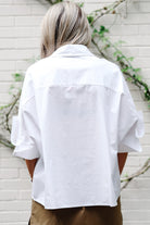 A person viewed from behind wearing an oversized white short-sleeve button-up shirt with rolled cuffs and tan pants, standing against a white brick wall with green vines.