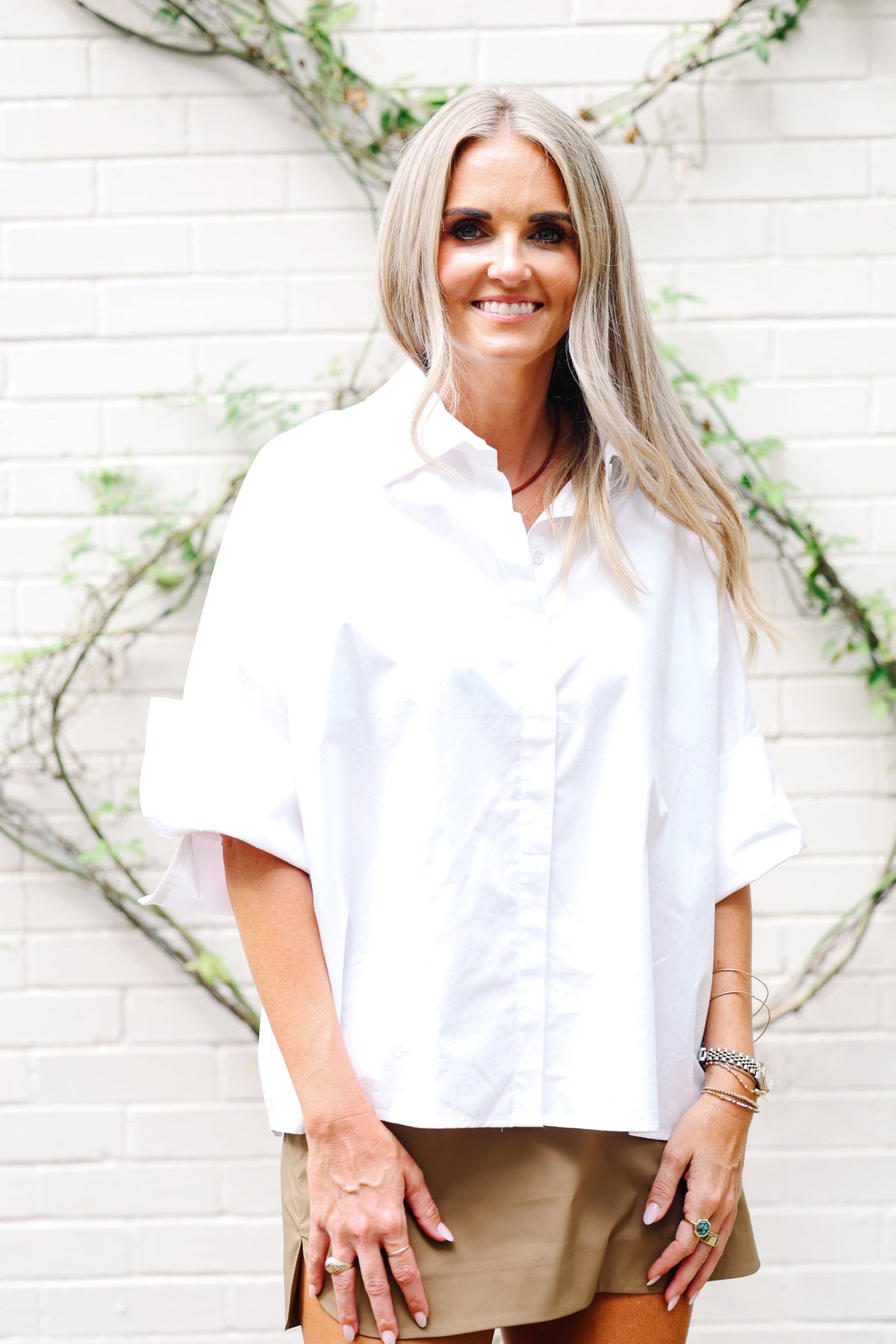 A woman with blonde hair wearing an oversized white short-sleeve button-down shirt and tan shorts, standing against a white brick wall with green vines.