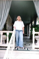 A woman stands on a porch wearing a white short-sleeve button-up shirt and light blue wide-leg jeans, framed by white railings and gray curtains.