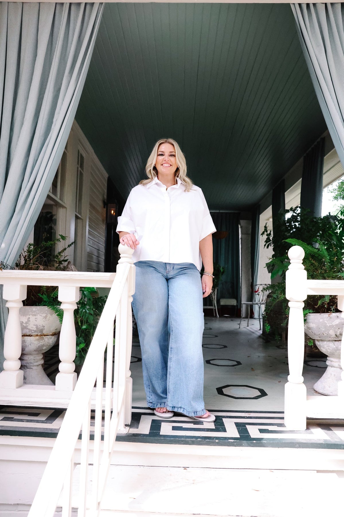 A woman stands on a porch wearing a white short-sleeve button-up shirt and light blue wide-leg jeans, framed by white railings and gray curtains.