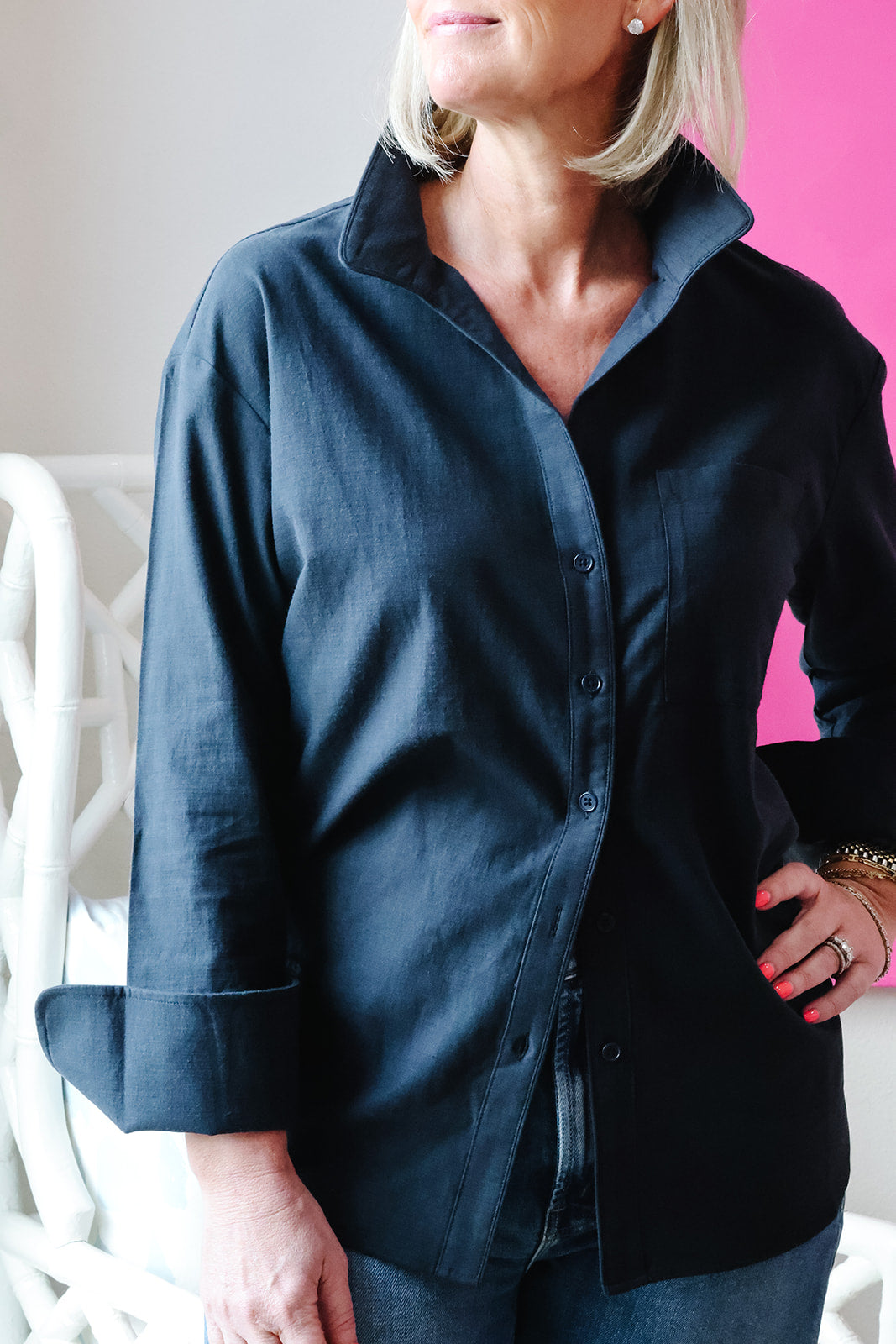 A woman wearing a dark navy blue button-down shirt with a collar and three-quarter sleeves, paired with blue jeans, standing against a split white and pink background.