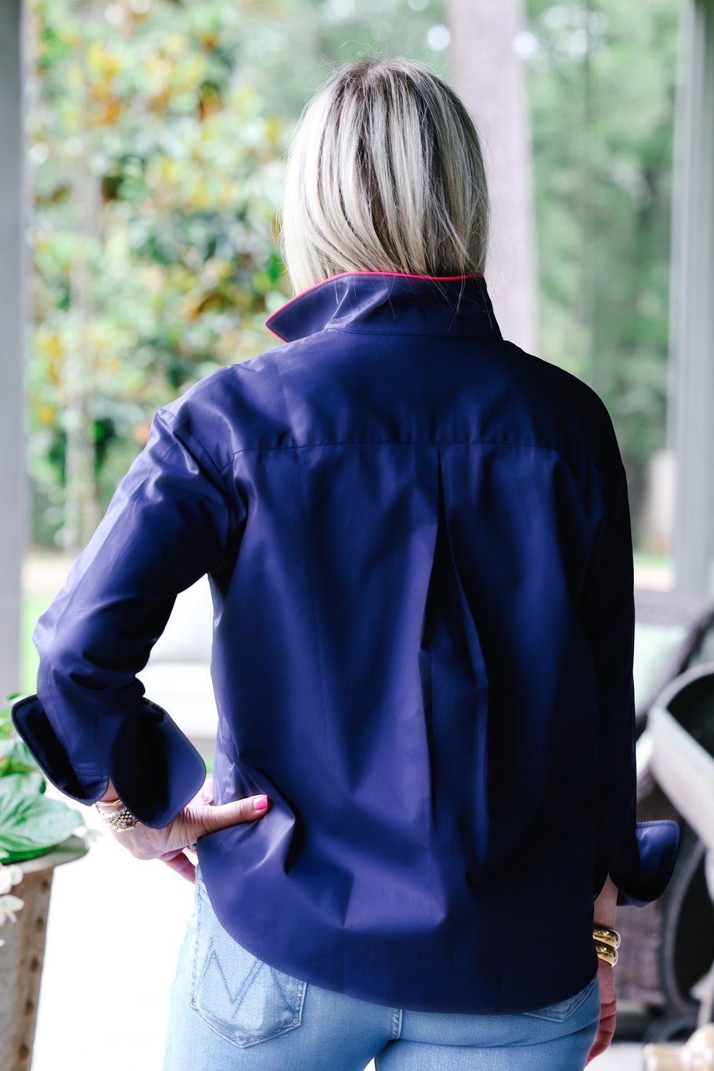 A woman viewed from behind wearing a navy blue shirt with a high collar and light blue jeans, standing on a porch with a blurred outdoor background.