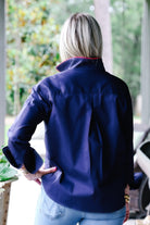 A woman viewed from behind wearing a navy blue shirt with a high collar and light blue jeans, standing on a porch with a blurred outdoor background.