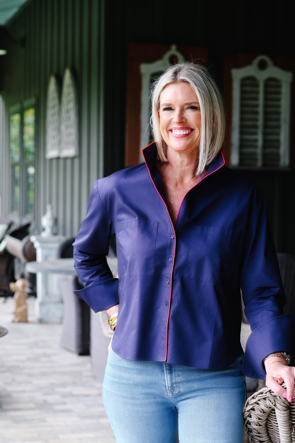 A smiling blonde woman wearing a navy blue button-up shirt with pink trim and light blue jeans, standing on a porch.
