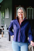 A smiling blonde woman wearing a navy blue button-up shirt with pink trim and light blue jeans, standing on a porch.