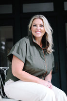 A smiling blonde woman sits on a white chair wearing an olive green short-sleeve button-up shirt with thin orange piping and white pants, posed in front of a dark doorway.