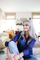 A smiling woman with long silver hair sits cross-legged on a light-colored sofa in a bright living room. She is wearing a dark navy blue satin pajama shirt with red piping and light blue jeans.