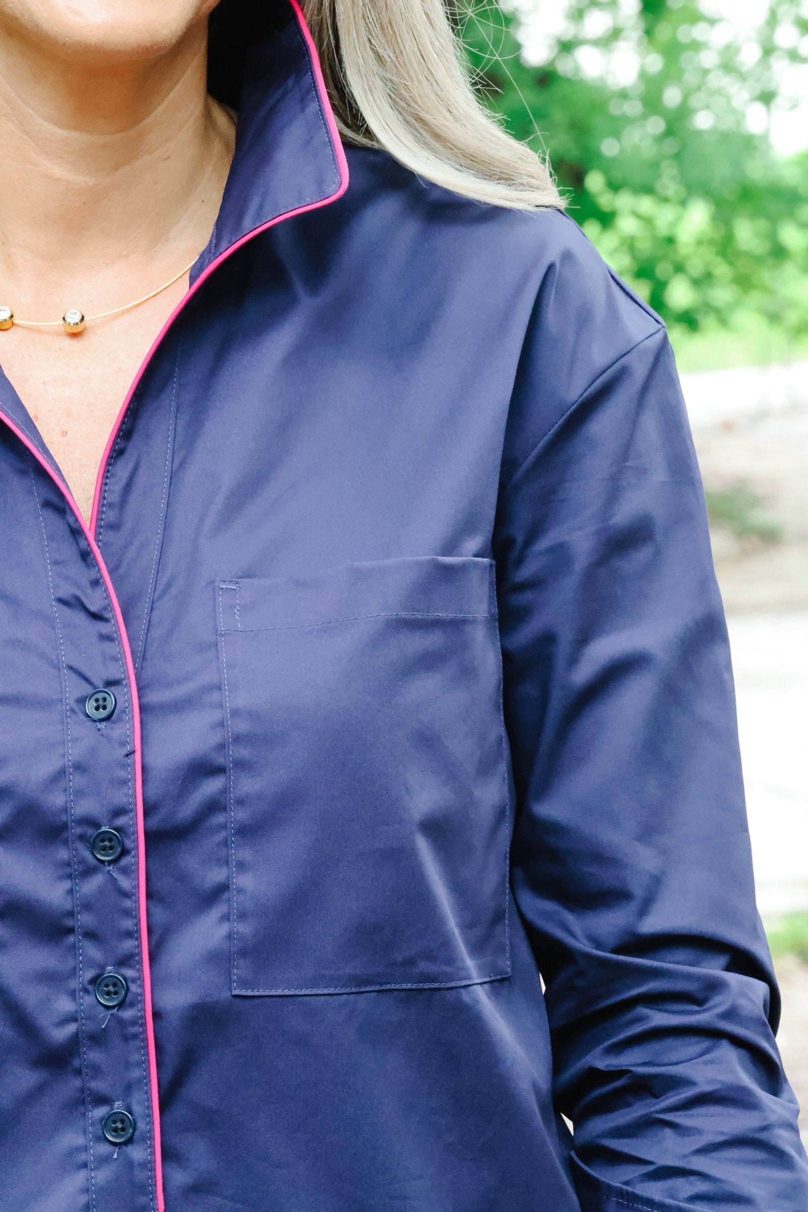 A close-up of a navy blue long-sleeve button-up shirt with bright pink piping trim along the collar, placket, and pocket, worn by a model outdoors.