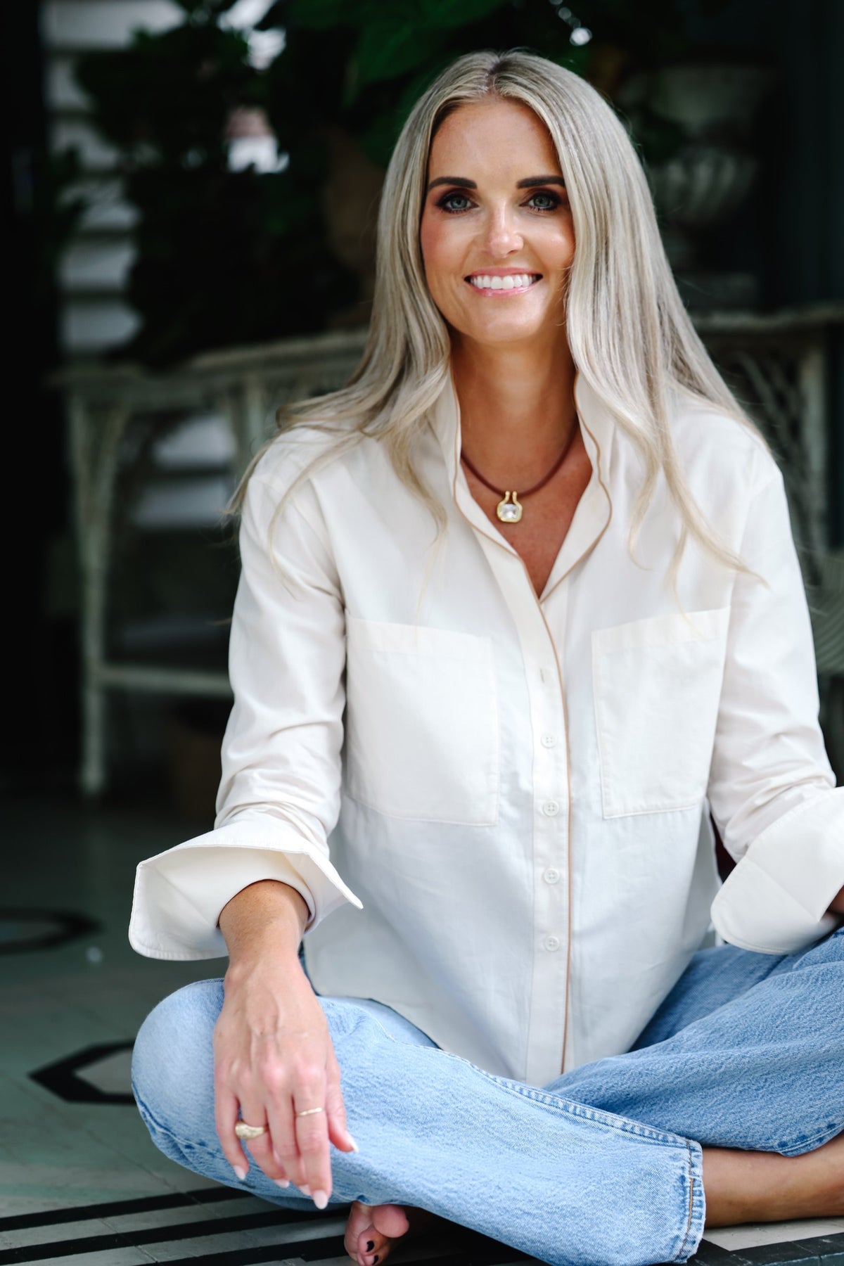 A smiling blonde woman sits cross-legged outdoors wearing a cream-colored long-sleeve button-down shirt and light blue jeans.